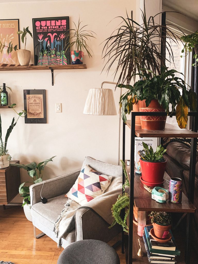 a picture of a corner of a room wth a cosy sitting chair with a blanket on it surrounded by art and house plants. A boho feel with cream, green and wood as the main colours